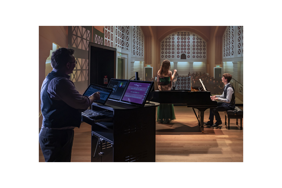 A man wearing a light blue shirt, using digital tablet to control a visual simulator, with two students, a female soprano and a male pianist, performing in front of a simulator.